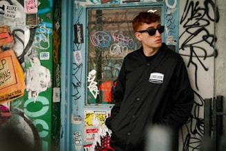 A person wearing sunglasses and a black jacket stands in an urban environment filled with colorful graffiti. The wall is covered in a variety of stickers, tags, and street art, contributing to a vibrant, artistic atmosphere.
