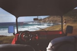 The view from inside a red vehicle, possibly a jeep, overlooking a coastal landscape. The dashboard and steering wheel are in the foreground, with side mirrors showing reflections of the ocean. In the background, a person stands near the edge of the coast, gazing at the waves crashing against the rocky shore. A hill rises to the right, and the sky is clear with minimal clouds.