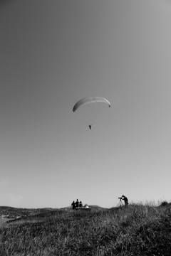 A paraglider is soaring in the sky while a group of people, some with a camera and tripod, are gathered on a grassy hill. The scene is captured in black and white, creating a serene and expansive atmosphere.