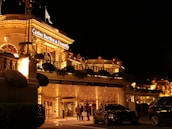 A luxurious casino at night, illuminated with warm, golden lights. The building features elegant architecture with columns and decorative elements. A few people are walking towards the entrance, and there are a couple of parked cars nearby.