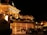 A luxurious casino at night, illuminated with warm, golden lights. The building features elegant architecture with columns and decorative elements. A few people are walking towards the entrance, and there are a couple of parked cars nearby.