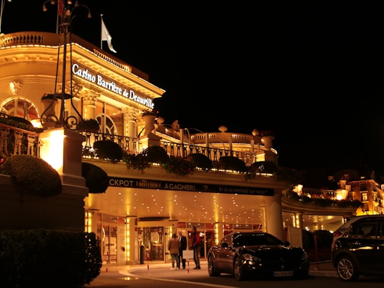 A luxurious casino at night, illuminated with warm, golden lights. The building features elegant architecture with columns and decorative elements. A few people are walking towards the entrance, and there are a couple of parked cars nearby.