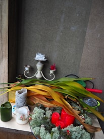 A decorative arrangement is placed against a textured wall, featuring a mix of dried and fresh leaves, various candles in different sizes and colors including red, white, and green, and a rustic candle holder. A round arrangement of red candles is set on a platter adorned with greenery and small decorative elements. The scene combines natural elements with crafted decorations, giving an eclectic and cozy appearance.