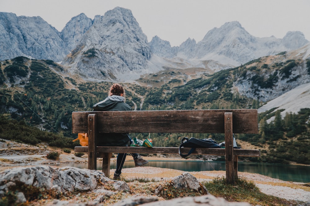 person wearing hooded jacket sitting on brown wooden bench near black bag facing on body of water viewing mountain during daytime