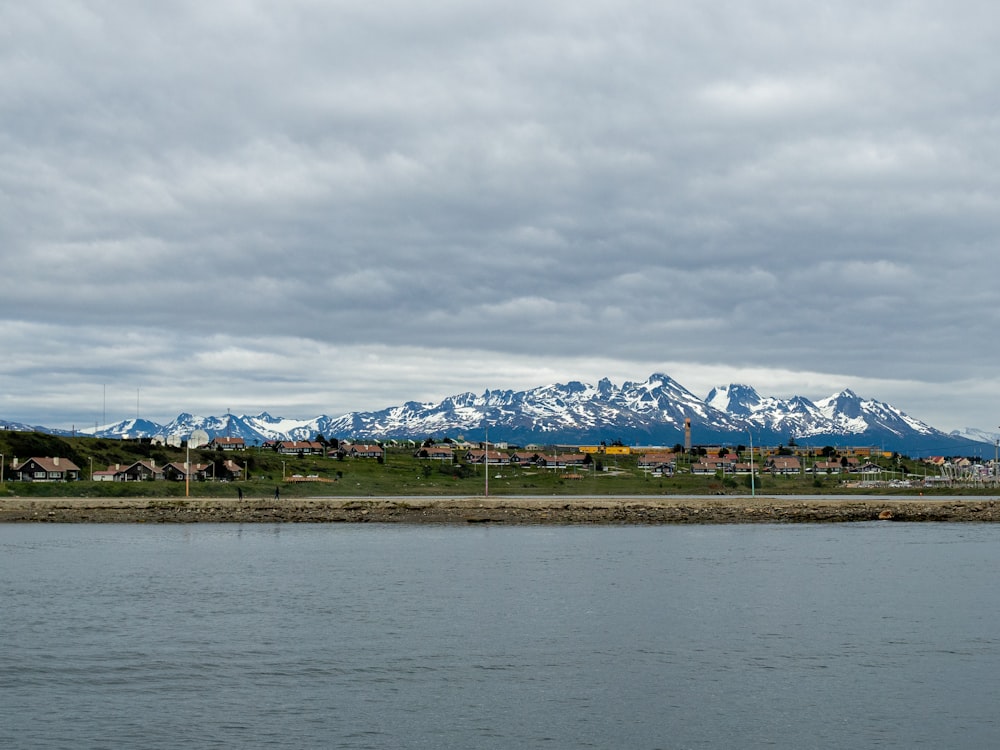 casas en campo verde cerca del cuerpo de agua que ve la montaña cubierta de nieve bajo el cielo blanco y gris