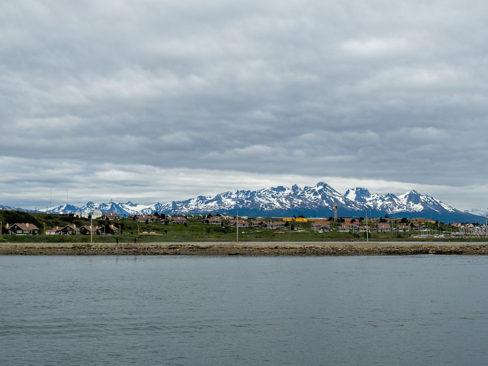 houses on green field near body of water viewing mountain covered with snow under white and gray sky