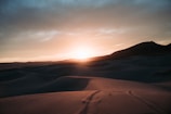 A vast desert landscape under a dramatic sunset, with rolling sand dunes and a dark mountain silhouette in the distance. The sky transitions from a bright, glowing horizon to darker, cloud-covered upper areas.