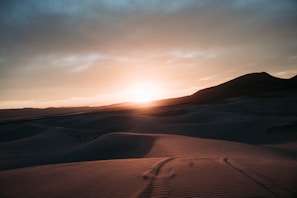 A vast desert landscape under a dramatic sunset, with rolling sand dunes and a dark mountain silhouette in the distance. The sky transitions from a bright, glowing horizon to darker, cloud-covered upper areas.