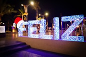 Brightly lit sign with the word 'FELIZ' is displayed against a nighttime backdrop. The letters are illuminated with blue and white lights, and the 'F' is decorated with a large red Santa hat. Streetlights and palm trees are visible in the background.