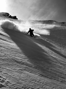 A skier is captured in mid-action, carving their way down a snowy mountain slope. The scene is dynamic, with a spray of snow trailing behind the skier, indicating high speed and skillful movement. The background is a mix of sky and rugged mountain terrain.