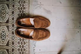 A pair of brown suede loafers is neatly placed on a wooden floor next to a patterned rug. The shoes appear to be well-crafted, with stitching details visible along the edges. The rug features intricate geometric patterns in shades of beige, brown, and black.
