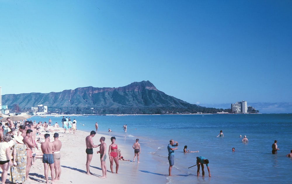people on beach viewing mountain and blue sea under blue and white sky