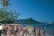 A crowded beach scene with numerous people in swimwear gathered around a yellow sign that reads 'Reef Coffee Shop.' In the background, there is a mountainous landscape and a blue ocean under a clear sky. Palm trees and lush greenery are visible, adding to the tropical atmosphere.