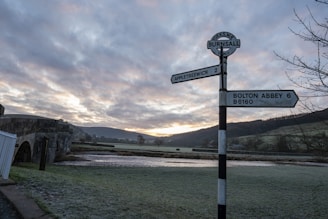 A rural scene with a road signpost in the foreground indicating directions to Appletreewick and Bolton Abbey. The background features a stone bridge crossing a river, with rolling hills and a cloudy sky at sunset.