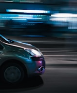 A car in motion is captured at night with blurred lights in the background, creating a sense of speed and movement. The lights reflect off the car's surface, adding vibrancy to the scene.