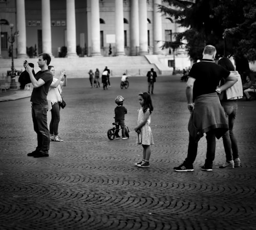In a public square, individuals engage in various activities, such as taking photos and walking. A little girl stands quietly while a child on a balance bike is nearby. The surroundings include tall columns and a broad open space.