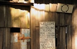 A wooden structure with a weathered appearance features a handmade sign promoting recycling. The sign, affixed to the wooden wall, contains text urging readers to follow instructions for effective recycling. Another smaller sign is visible with a recycling symbol above the larger sign. The setting gives an impression of simplicity and a focus on eco-friendly practices.
