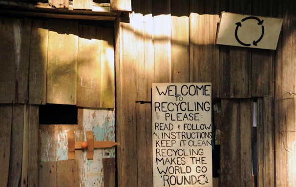 A wooden structure with a weathered appearance features a handmade sign promoting recycling. The sign, affixed to the wooden wall, contains text urging readers to follow instructions for effective recycling. Another smaller sign is visible with a recycling symbol above the larger sign. The setting gives an impression of simplicity and a focus on eco-friendly practices.