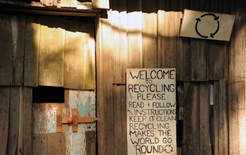 A wooden structure with a weathered appearance features a handmade sign promoting recycling. The sign, affixed to the wooden wall, contains text urging readers to follow instructions for effective recycling. Another smaller sign is visible with a recycling symbol above the larger sign. The setting gives an impression of simplicity and a focus on eco-friendly practices.