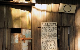 A wooden structure with a weathered appearance features a handmade sign promoting recycling. The sign, affixed to the wooden wall, contains text urging readers to follow instructions for effective recycling. Another smaller sign is visible with a recycling symbol above the larger sign. The setting gives an impression of simplicity and a focus on eco-friendly practices.