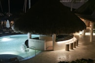 A tropical-themed lounge area features a thatched roof bar surrounded by a circular pool with gentle lighting. Straw stools line the curved bar counter, and stone pathways lead through the layout. The ambient, relaxed night scene gives off an inviting feel.