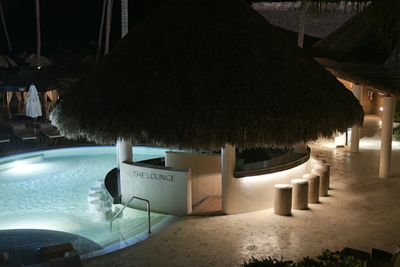 A tropical-themed lounge area features a thatched roof bar surrounded by a circular pool with gentle lighting. Straw stools line the curved bar counter, and stone pathways lead through the layout. The ambient, relaxed night scene gives off an inviting feel.