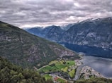 A scenic view of a deep blue fjord surrounded by steep, green mountains under a cloudy sky. In the foreground, a small village with fields and roads can be seen along the water's edge. Snow-capped mountains are visible in the distance.
