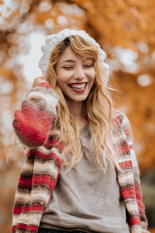 A smiling person wrapped in a thick knit cardigan, standing outside on a crisp autumn day.