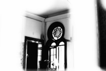 A monochrome photograph of an interior space featuring a window with decorative circular and semi-circular designs above it. The room has high ceilings with visible molding and the walls seem to be adorned with ornate tiling near the bottom. Light streams in from the window, creating a high contrast effect.