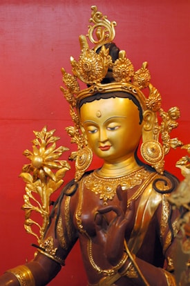 A golden statue of a deity adorned with intricate ornaments and a crown, set against a rich red background. The figure holds a scepter topped with a floral design and is depicted in serene meditation, with finely detailed carvings and patterns.