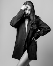 A black and white photograph of a person with long hair wearing an oversized blazer. The individual has one hand placed on their forehead, and the blazer partially conceals their body.