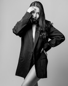 A black and white photograph of a person with long hair wearing an oversized blazer. The individual has one hand placed on their forehead, and the blazer partially conceals their body.