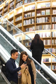 Two people are on an escalator inside a bookstore. They appear to be engaged with something on a smartphone, with a backdrop of multiple shelves filled with books. Another person is ascending on the parallel escalator.