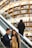 Two people are on an escalator inside a bookstore. They appear to be engaged with something on a smartphone, with a backdrop of multiple shelves filled with books. Another person is ascending on the parallel escalator.