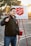 A person is smiling and holding a bell, standing in front of a Salvation Army donation setup with a sign and a red kettle. The background features a parking lot with trees and a setting sun, casting a warm glow.
