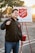 A person is smiling and holding a bell, standing in front of a Salvation Army donation setup with a sign and a red kettle. The background features a parking lot with trees and a setting sun, casting a warm glow.
