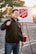 A person is smiling and holding a bell, standing in front of a Salvation Army donation setup with a sign and a red kettle. The background features a parking lot with trees and a setting sun, casting a warm glow.