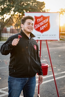 A person is smiling and holding a bell, standing in front of a Salvation Army donation setup with a sign and a red kettle. The background features a parking lot with trees and a setting sun, casting a warm glow.