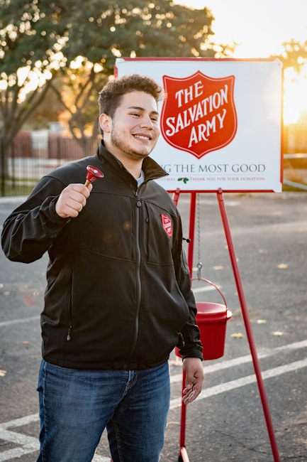 A person is smiling and holding a bell, standing in front of a Salvation Army donation setup with a sign and a red kettle. The background features a parking lot with trees and a setting sun, casting a warm glow.