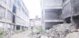 A dilapidated urban environment featuring large abandoned concrete buildings with extensive graffiti on the walls. The ground is strewn with rubble and debris, suggesting a site of neglect and decay. Some plants are visible among the ruins, indicating a slight return of nature.