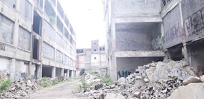 A dilapidated urban environment featuring large abandoned concrete buildings with extensive graffiti on the walls. The ground is strewn with rubble and debris, suggesting a site of neglect and decay. Some plants are visible among the ruins, indicating a slight return of nature.