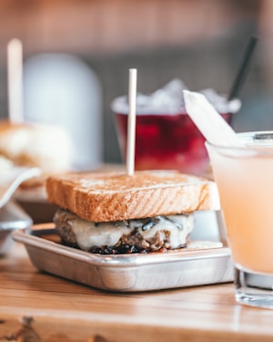 A close-up of a gourmet sandwich featuring toasted bread and a savory filling, placed in a small metal tray. The background includes a glass of red beverage with ice and another glass holding a pale peach-colored drink. The focus is sharp on the sandwich, with the drinks slightly out of focus.