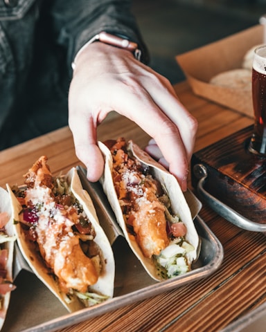 A hand reaches for a tray of tacos filled with crispy fried fish, topped with fresh salsa and a sprinkle of cheese. The scene also features a table set with a small glass of beer, suggesting a relaxed dining atmosphere.