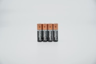 Four AA batteries are standing upright in a straight line. The batteries have a copper-colored top half and a black bottom half, with branding and text visible on the black section. They are placed against a plain, light-colored background.