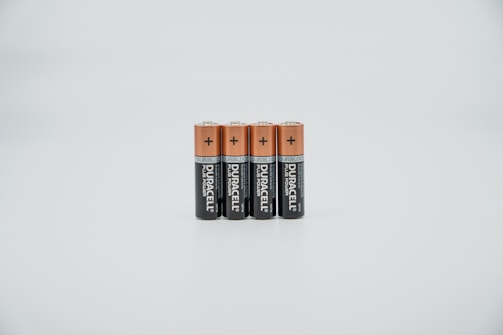 Four AA batteries are standing upright in a straight line. The batteries have a copper-colored top half and a black bottom half, with branding and text visible on the black section. They are placed against a plain, light-colored background.