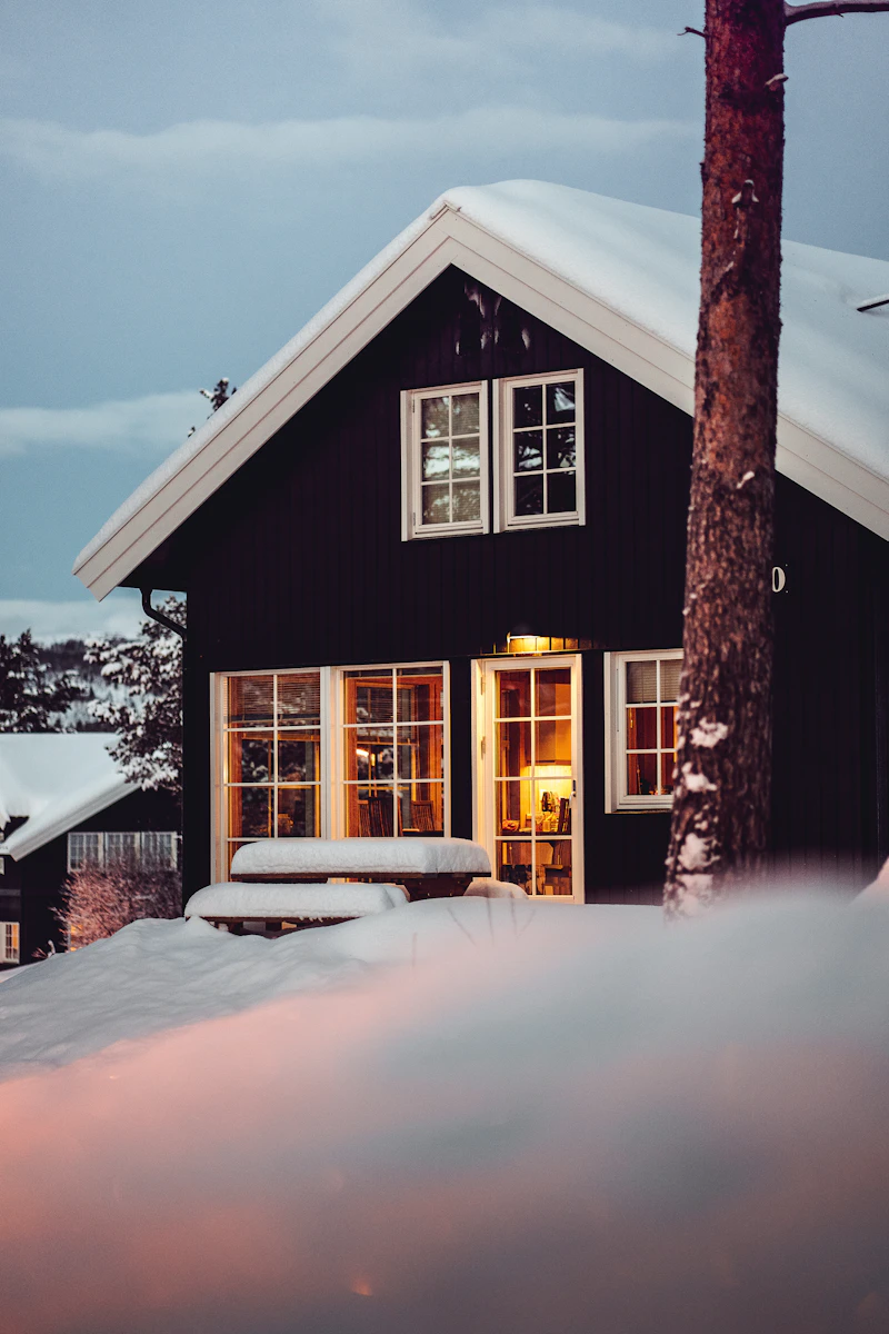 A dark wood house with lit windows at dusk, snow on its roof