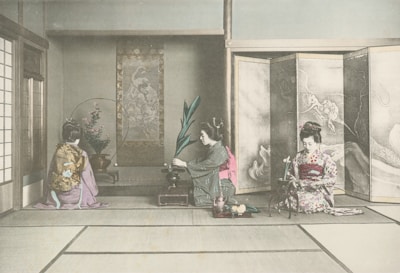 Three women dressed in traditional kimono are engaged in a cultural activity inside a room with tatami flooring. One is arranging flowers, while the other two appear to be observing or participating in a traditional Japanese tea ceremony. The room features classical Japanese art and decorations, including a painted screen and hanging scroll.