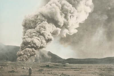 A large plume of smoke and ash rises from the ground, indicating volcanic activity. The landscape appears barren with rocky terrain, and there are mountains in the background. A solitary figure is seen walking toward the volcano.