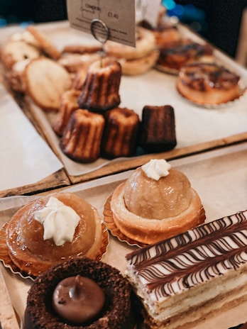 An assortment of pastries is displayed on a wooden tray. There are two round pastries topped with whipped cream, a chocolate cake slice with icing patterns, and various baked goods like Canelés and tarts in the background.