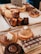 An assortment of pastries is displayed on a wooden tray. There are two round pastries topped with whipped cream, a chocolate cake slice with icing patterns, and various baked goods like Canelés and tarts in the background.